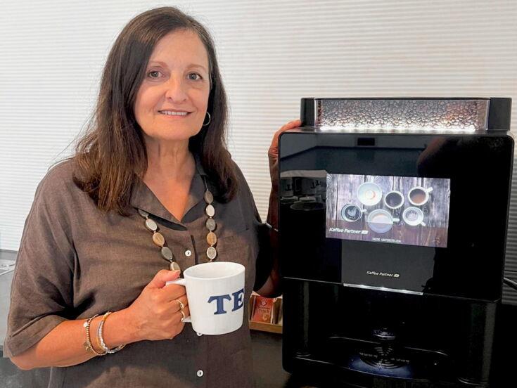 Andrea Lang von der Firma Texa steht lächelnd, mit einer Tasse in der Hand, neben einem Kaffeevollautomaten von Kaffee Partner